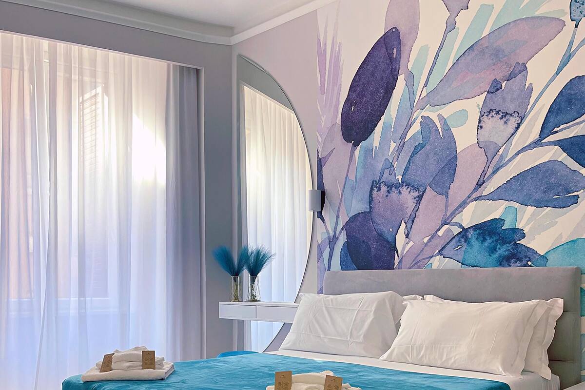 Ein modernes Schlafzimmer mit einem großen Bett, einer dekorativen Wandmalerei und Blick ins Freie durch transparente Vorhänge.