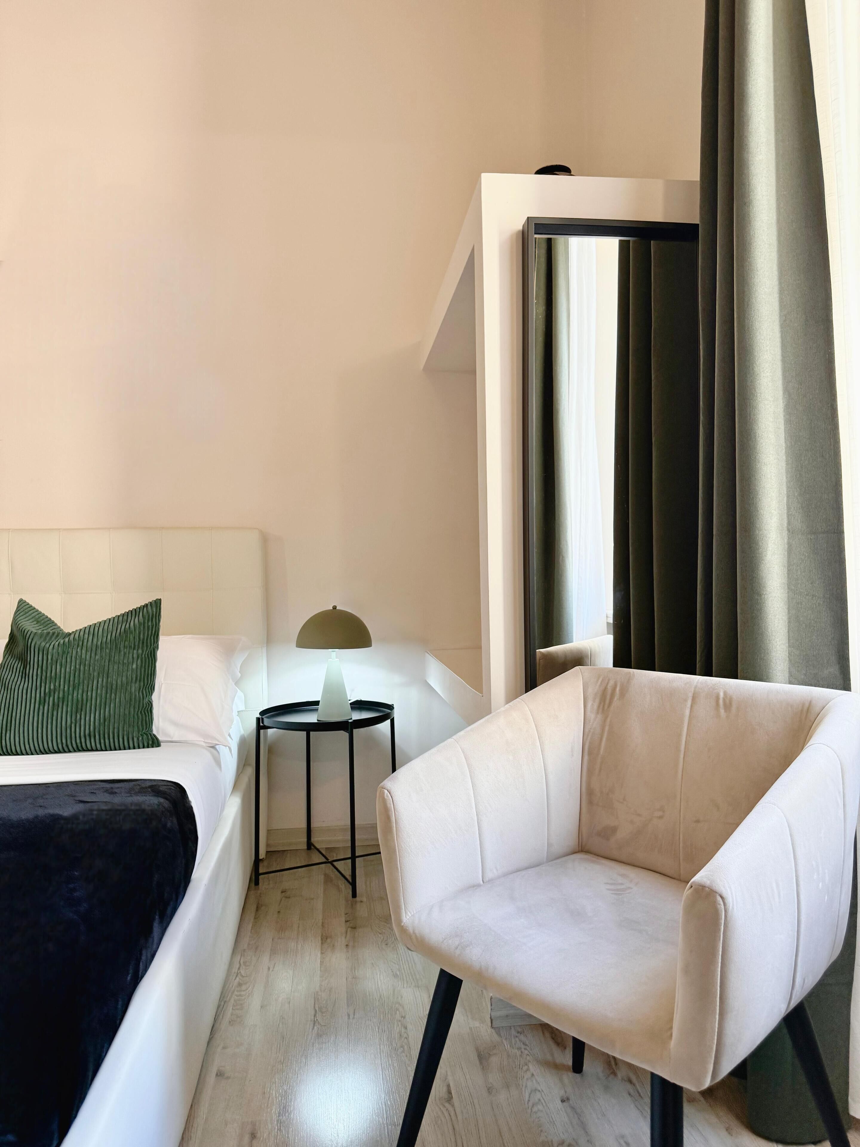 A hotel room with a bed, a chair, a small table with a lamp, and a window with curtains.