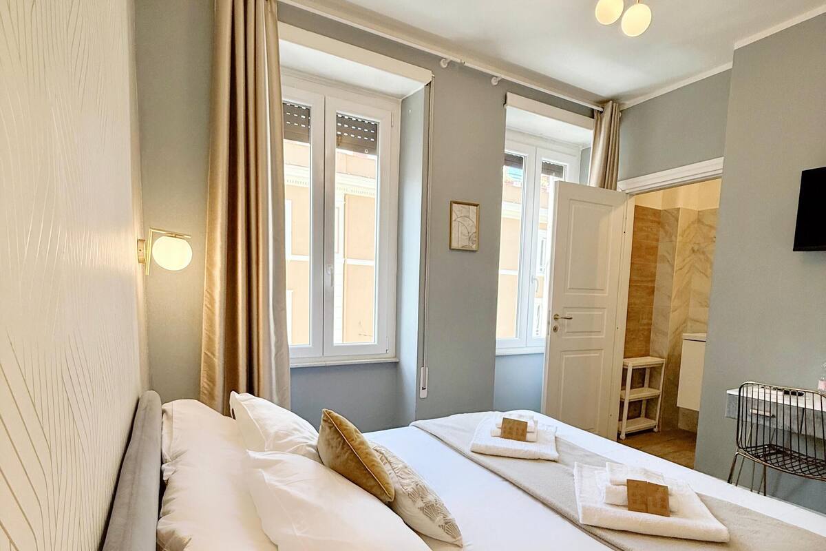 Ein ordentlich bezogenes Bett mit weißen Leinentüchern und einem einzelnen Zierkissen, ein Nachttisch mit einer kleinen Lampe und ein Fenster mit Vorhängen.