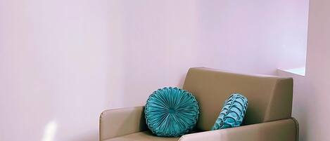 Un fauteuil beige agrémenté de deux coussins à motifs, un pouf bleu et trois tableaux abstraits accrochés à un mur rose pâle.