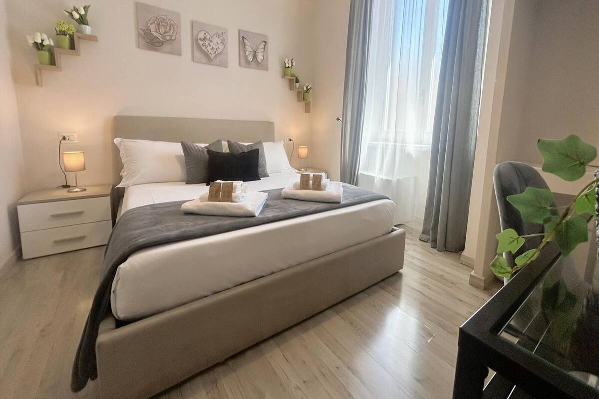 Ein modernes Schlafzimmer mit einem großen Bett, Nachttischen mit Lampen, Wandbildern und Blick ins Freie durch transparente Vorhänge.