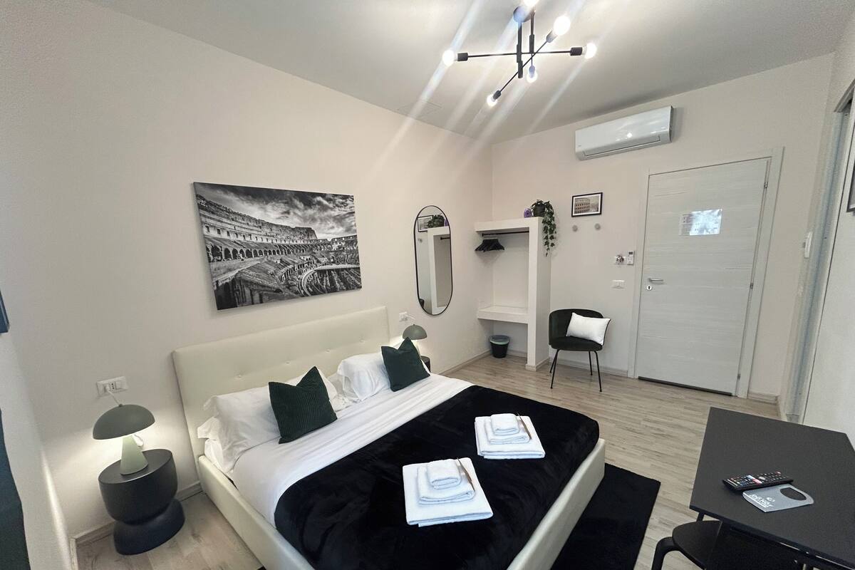 Ein modernes Hotelzimmer mit einem großen Bett, einem Schreibtisch, einem Stuhl, einem Spiegel und einem Bild von einem antiken Amphitheater.