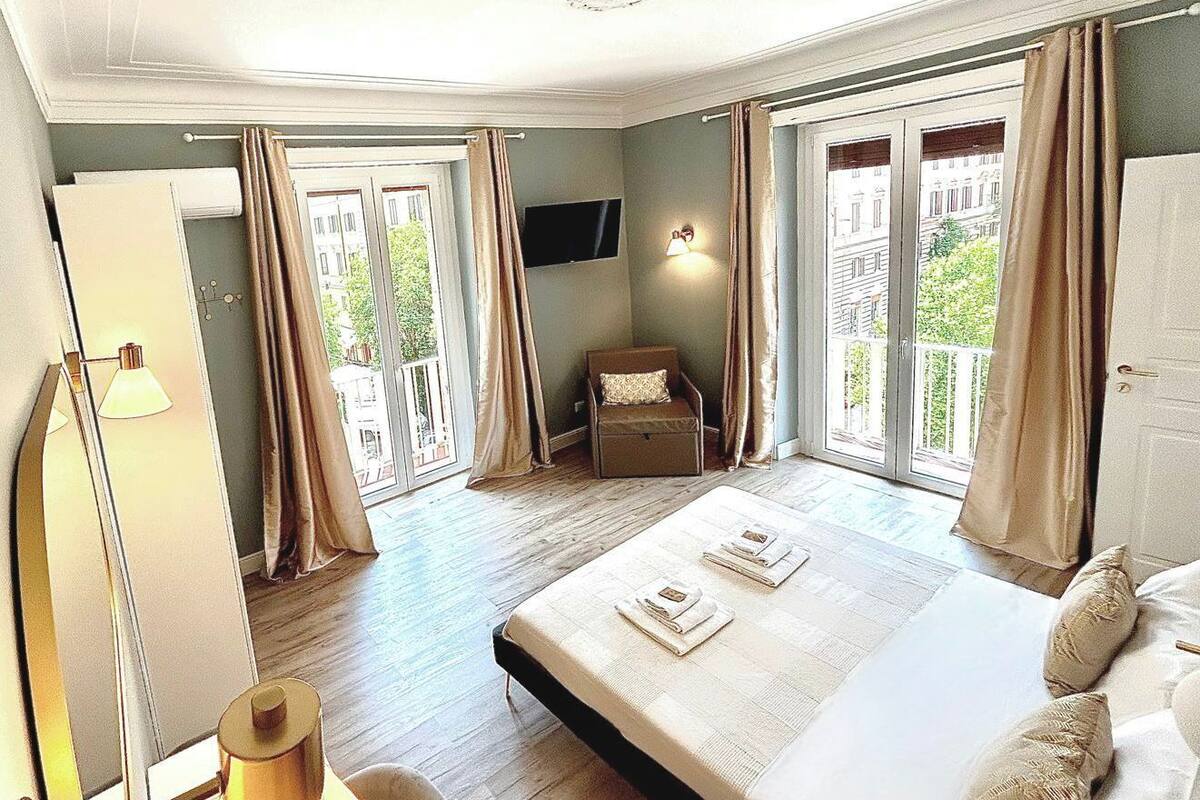 Ein Hotelzimmer mit einem großen Bett, einem Sessel, einem Schreibtisch mit Lampe und einem an der Wand befestigten Fernseher.