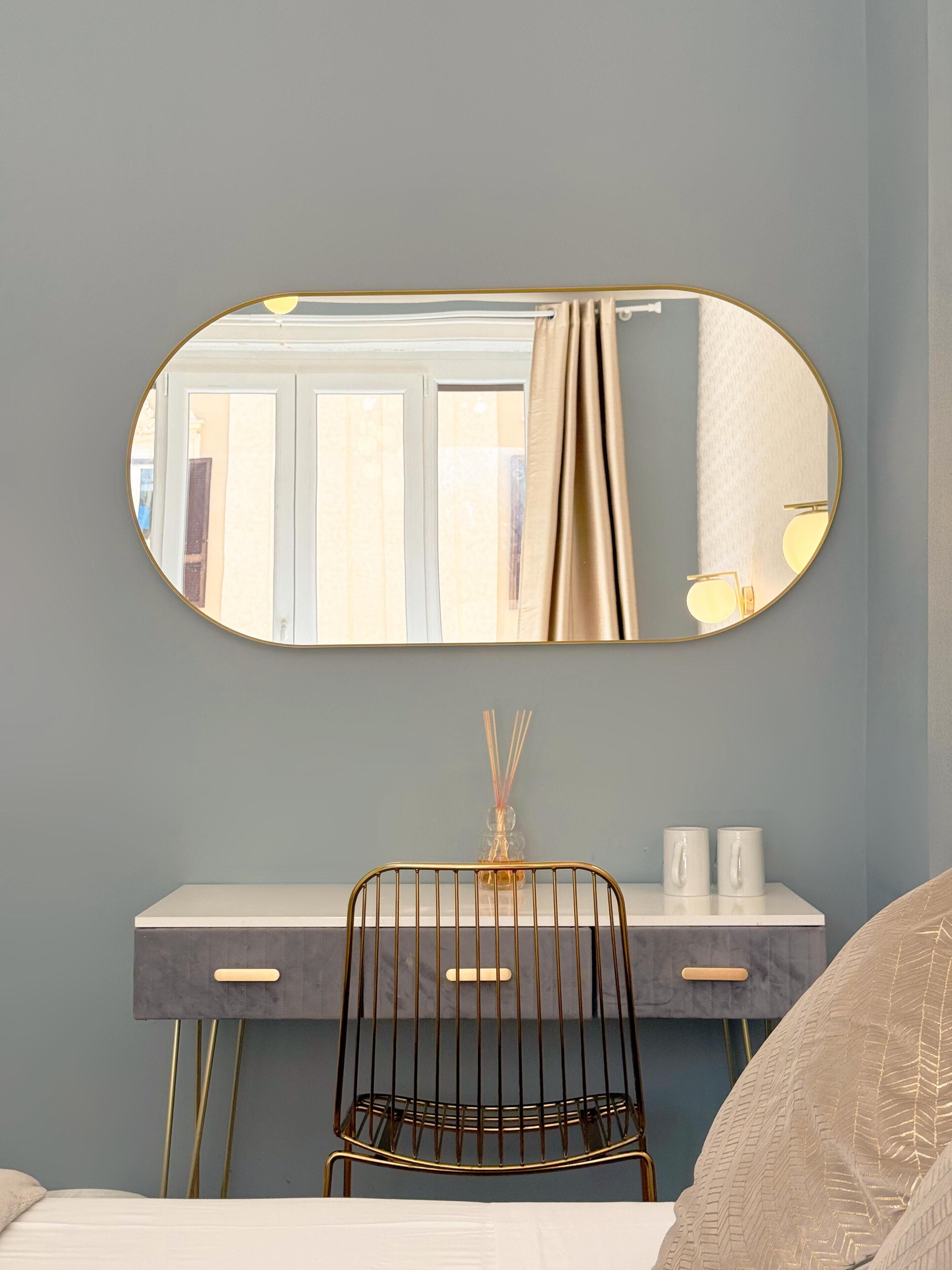 Un dormitorio con cama, un escritorio con silla, un espejo redondo y una ventana con cortinas.