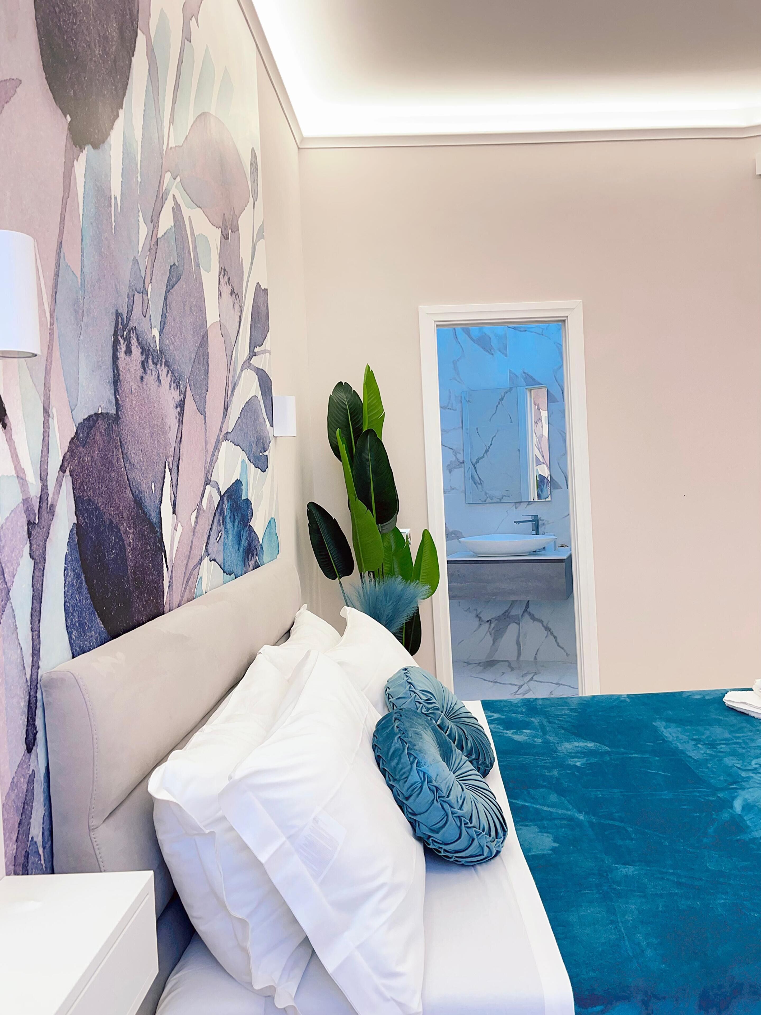 A bedroom with a bed, a large abstract painting, a potted plant, and a view of a bathroom through an open door.