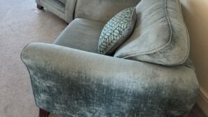 Een grijze bank met gekleurde kussens in een kamer met een grijze deur, een aan de muur bevestigd bordje met de tekst 'BACH' en een lijst met een afbeelding.