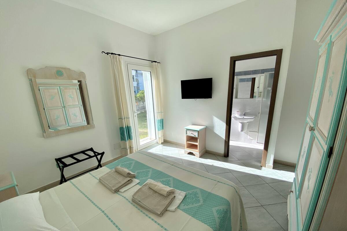 A bedroom with a bed, a small bedside table, a TV, and a bathroom visible through an open door.