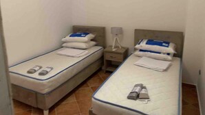 Quarto de hotel pequeno e individual com duas camas de solteiro, cada uma com travesseiros e cobertor. Há uma mesa de cabeceira com uma luminária e um telefone.