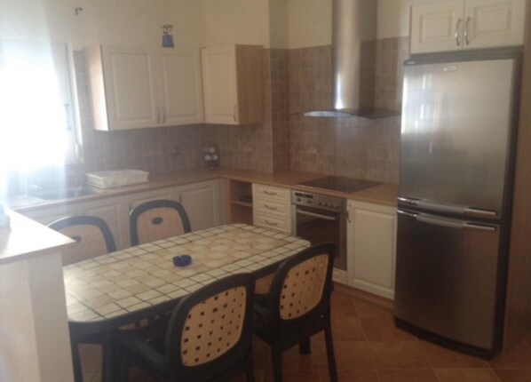 A compact kitchen with a refrigerator, stove, and sink. There are two chairs and a table with a tiled top.