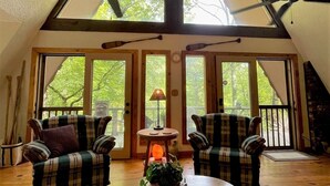 A cozy room with a high ceiling, wooden beams, plaid furniture, a central table with a lamp, and large windows offering a view of greenery outside. - Granny Squirrel Getaway (Andrews)