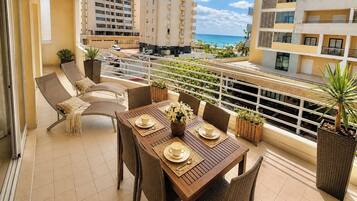 A balcony with a dining table set for four, surrounded by wicker chairs, with a view of a beach and high-rise buildings.