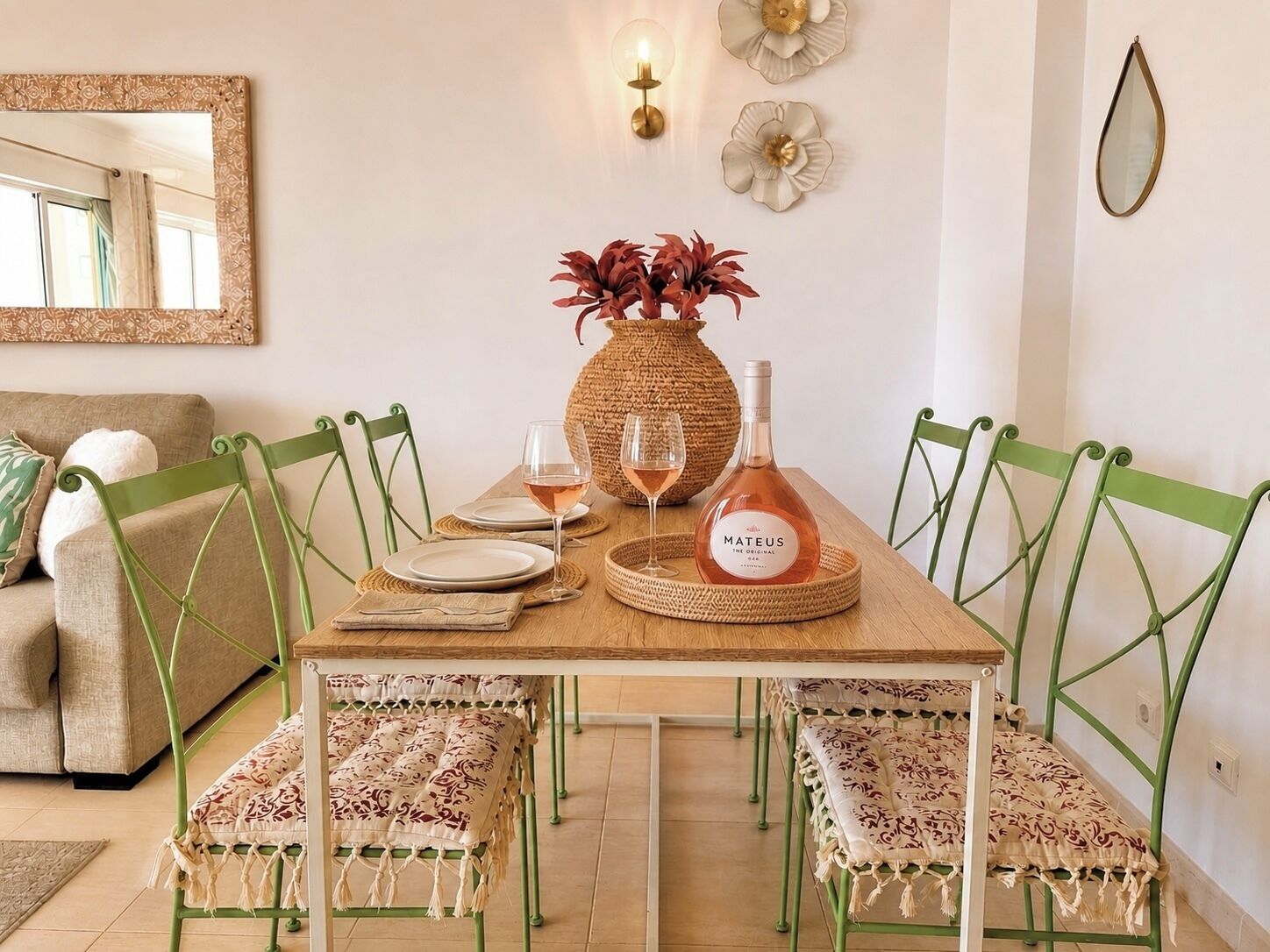 A dining area with a wooden table, green chairs, a woven vase with red flowers, two wine glasses, and a bottle of Mateus wine.