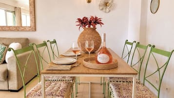 A dining area with a wooden table, green chairs, a woven vase with red flowers, two wine glasses, and a bottle of Mateus wine.