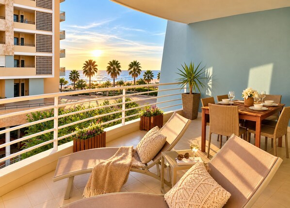 A balcony with a dining set, lounge chairs, and a view of palm trees and the sea.