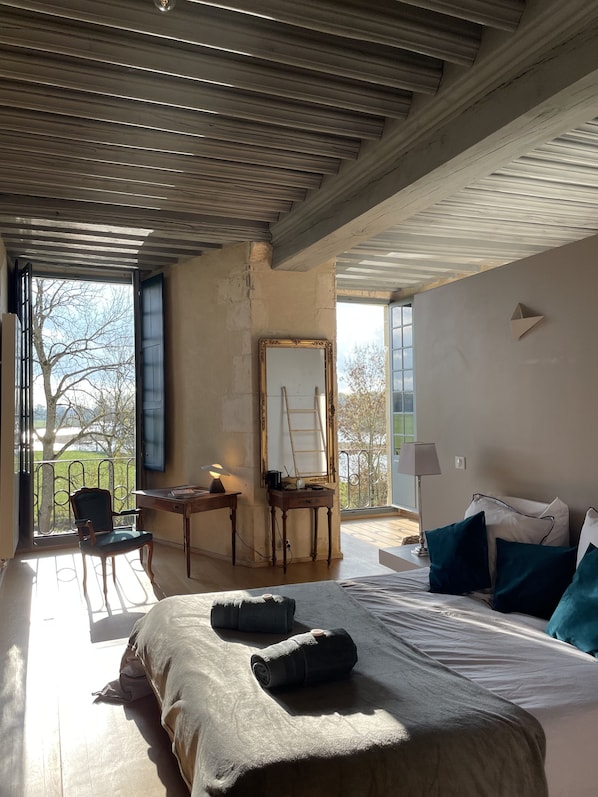 A bedroom with a large bed, a desk, a chair, a mirror, and a balcony with a view of trees. - Château de Meauce (Saincaize-Meauce)