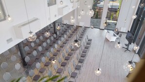 Et rummeligt mødelokale med rækker af stole og et hvidt bord, moderne pendellamper og udsigt gennem store glasdøre.