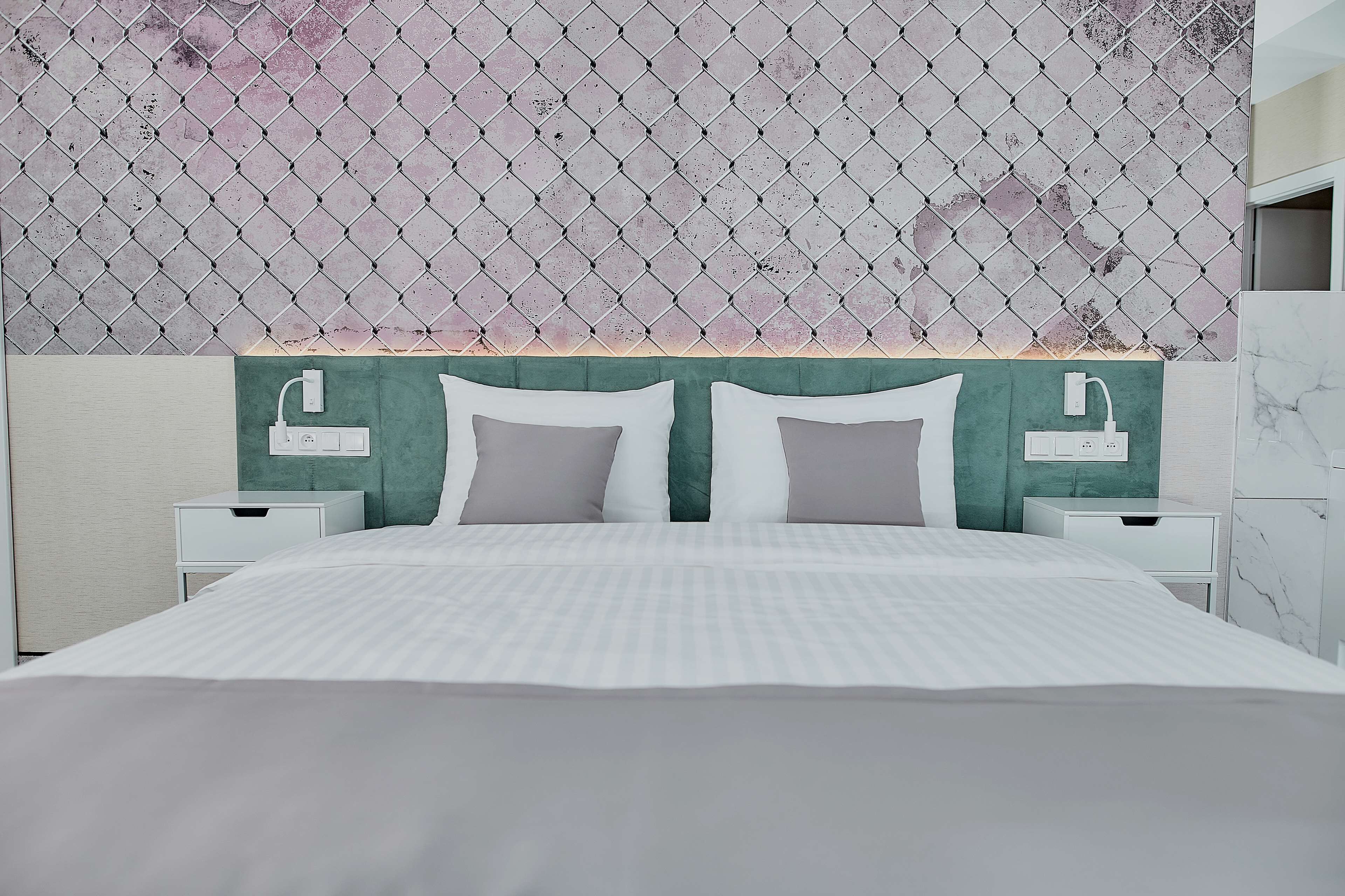 Un lit bien fait, recouvert d’une couette à rayures blanches et grises, deux tables de chevet avec des lampes, et un mur orné d’un motif en losanges.