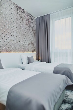 Une chambre d’hôtel moderne avec deux lits, une grande fenêtre avec des rideaux légers, un mur décoratif et une table de chevet avec une lampe.