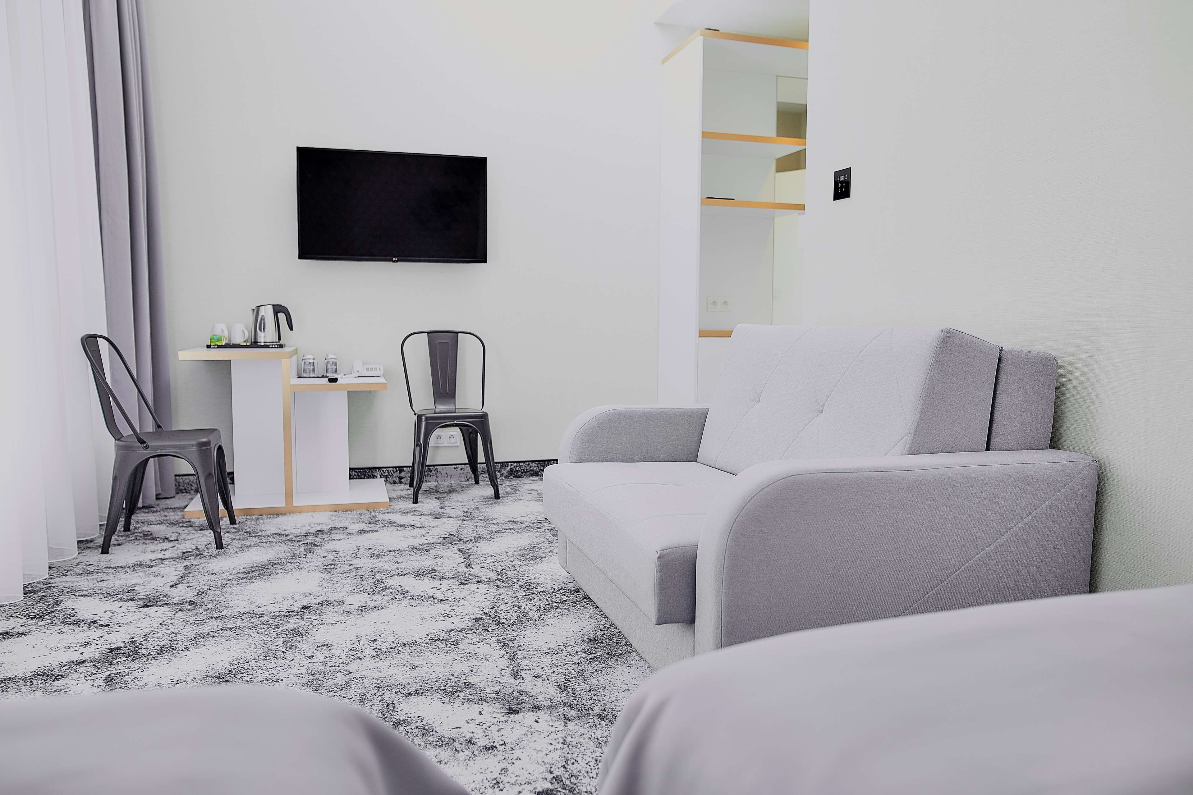 A modern hotel room with a grey sofa, a small desk with a kettle, and a TV mounted on the wall.