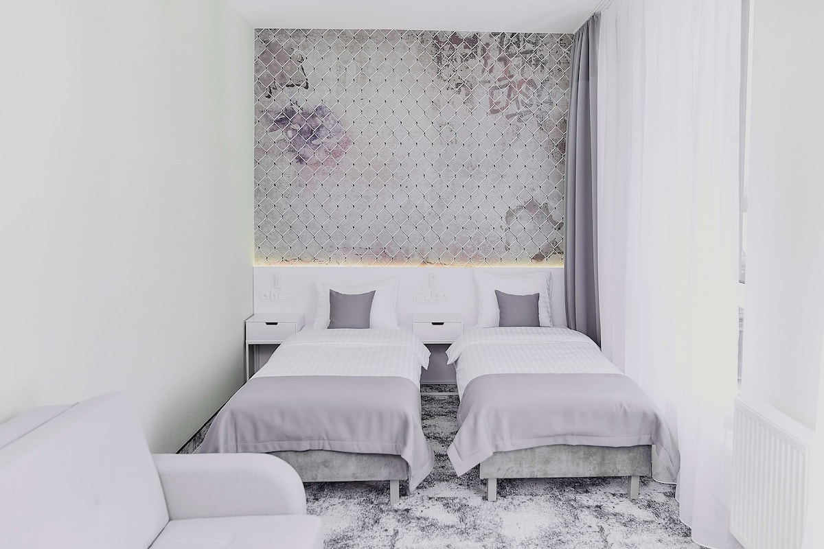 A modern hotel room with two single beds, a large window with curtains, a white wall, and a textured artwork above the beds.