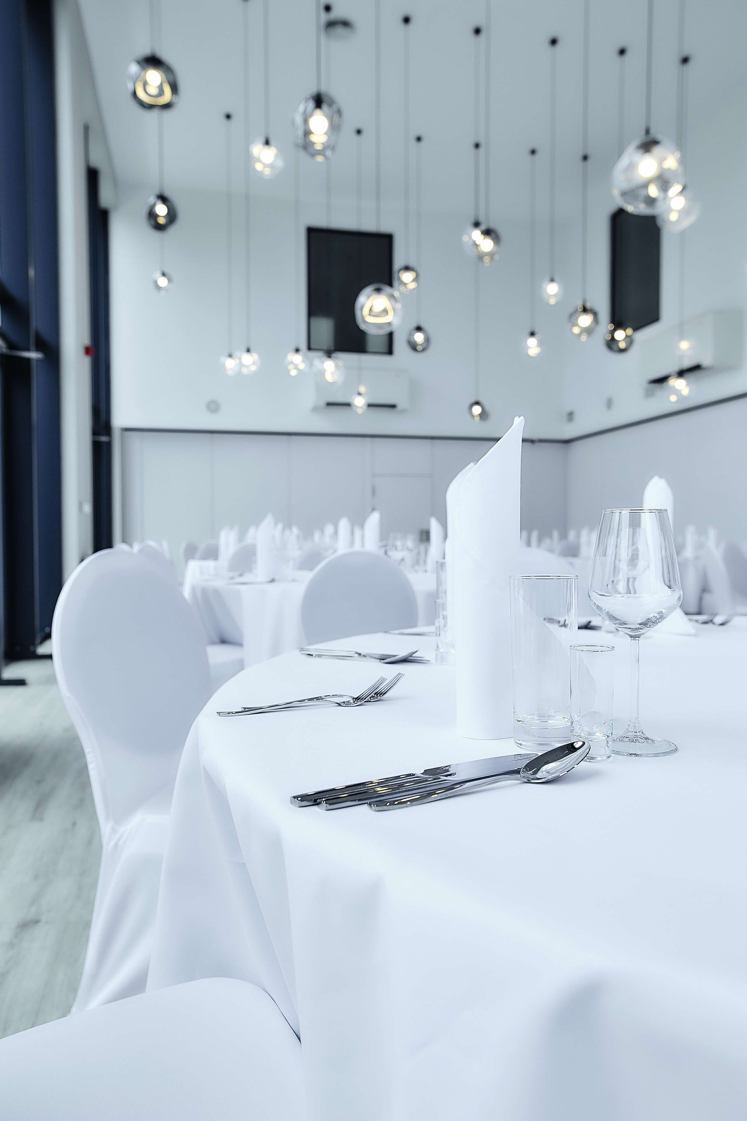 A banquet hall with round tables, white tablecloths, and hanging pendant lights.