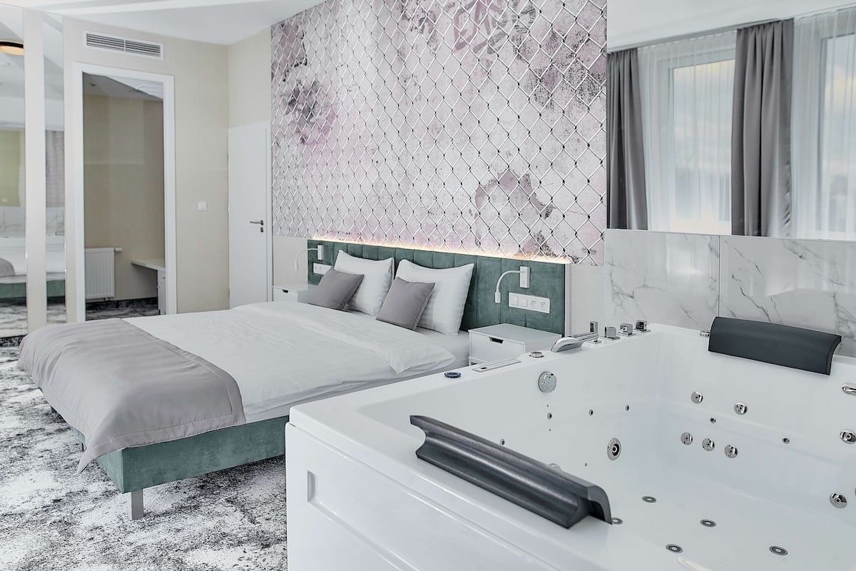 A modern hotel room with a large bed, a separate sitting area, a bathtub, and a view of the city.