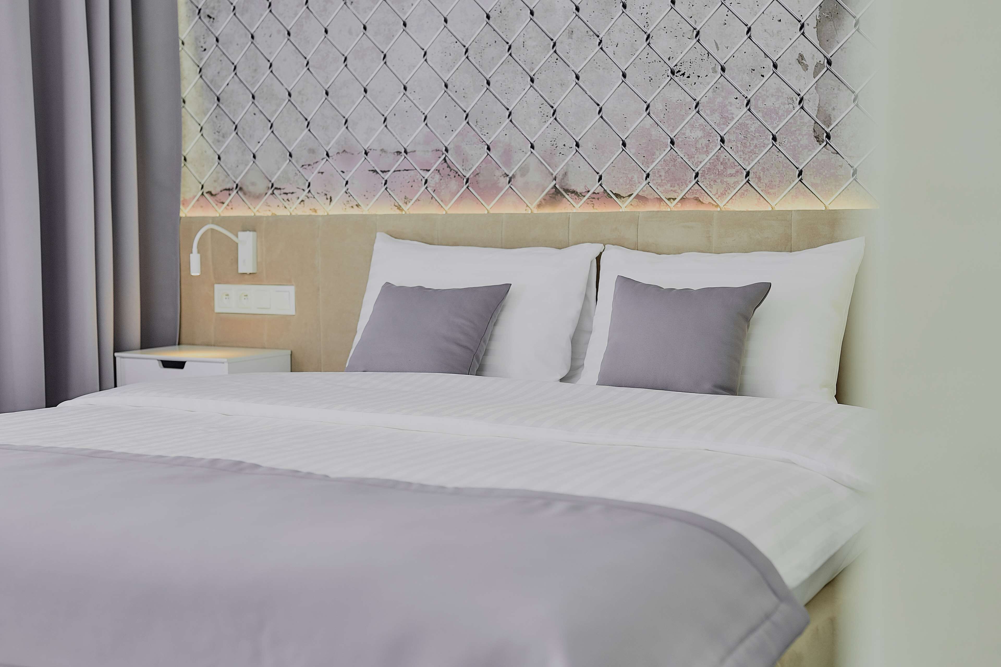 Una cama bien hecha con ropa de cama blanca y gris, un cabecero con diseño de rombos y una mesita de noche con una lámpara blanca y toma de corriente.
