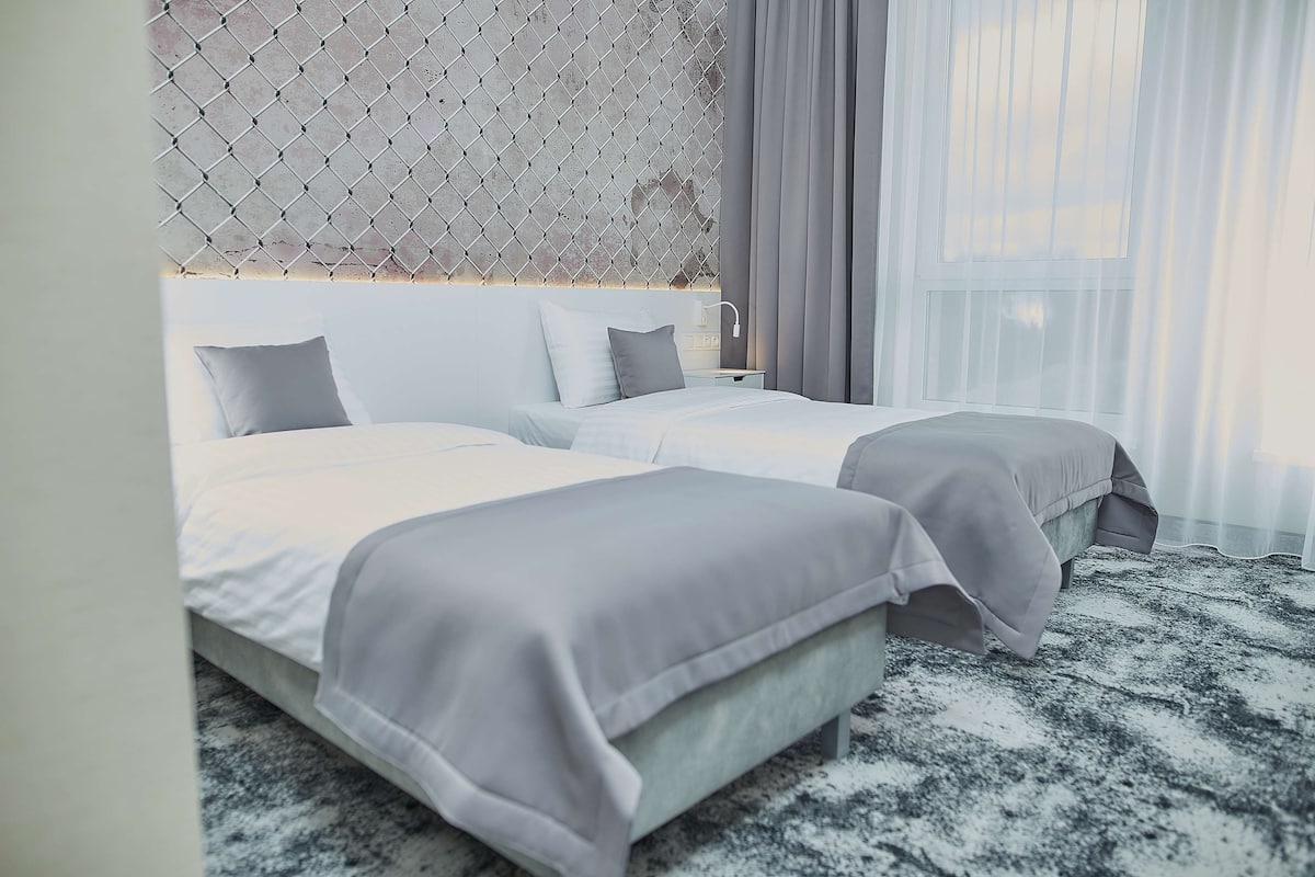 A double bed with a grey headboard and two pillows, a bedside table with a lamp, and a window with sheer curtains.