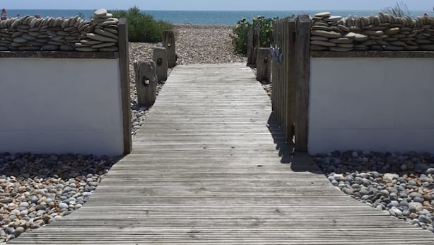 Une promenade en bois menant à une plage, bordée de murs de pierre et de galets.