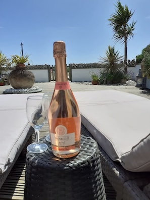 A bottle of Prosecco Rosé on a wicker table with a glass, set on a sunlit outdoor lounge area with cushions and a view of a coastal landscape. - Ocean Front Beach House With Direct Beach Access (Pevensey)