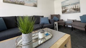 Un salon moderne avec un canapé gris, une table basse et deux fauteuils. Deux tableaux ornent les murs, une plante en pot est posée sur la table basse, et un plateau contient une bougie et des bâtonnets d’encens.