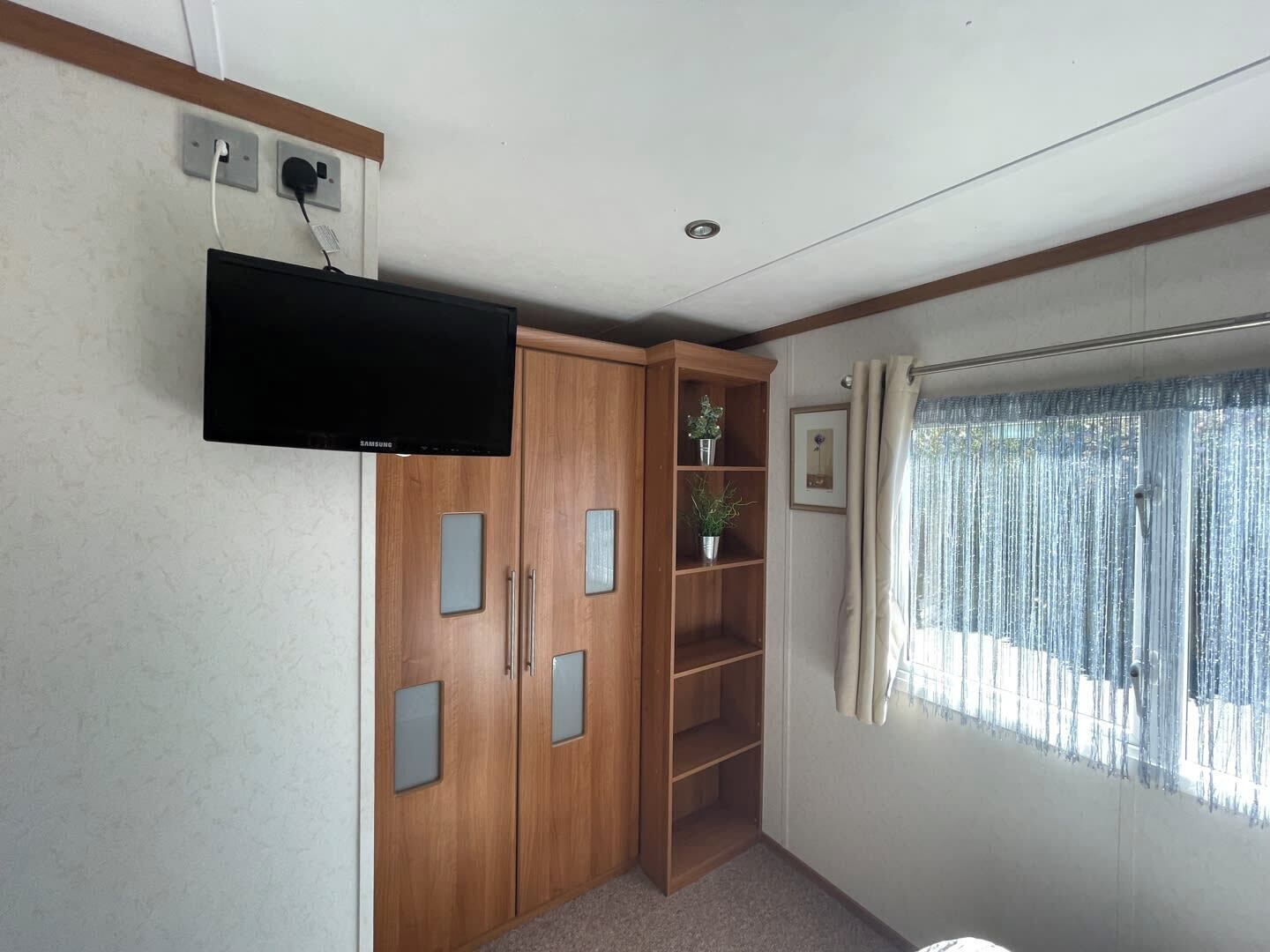 A room with a wooden wardrobe, a mounted television, a window with curtains, and a small plant on a shelf.