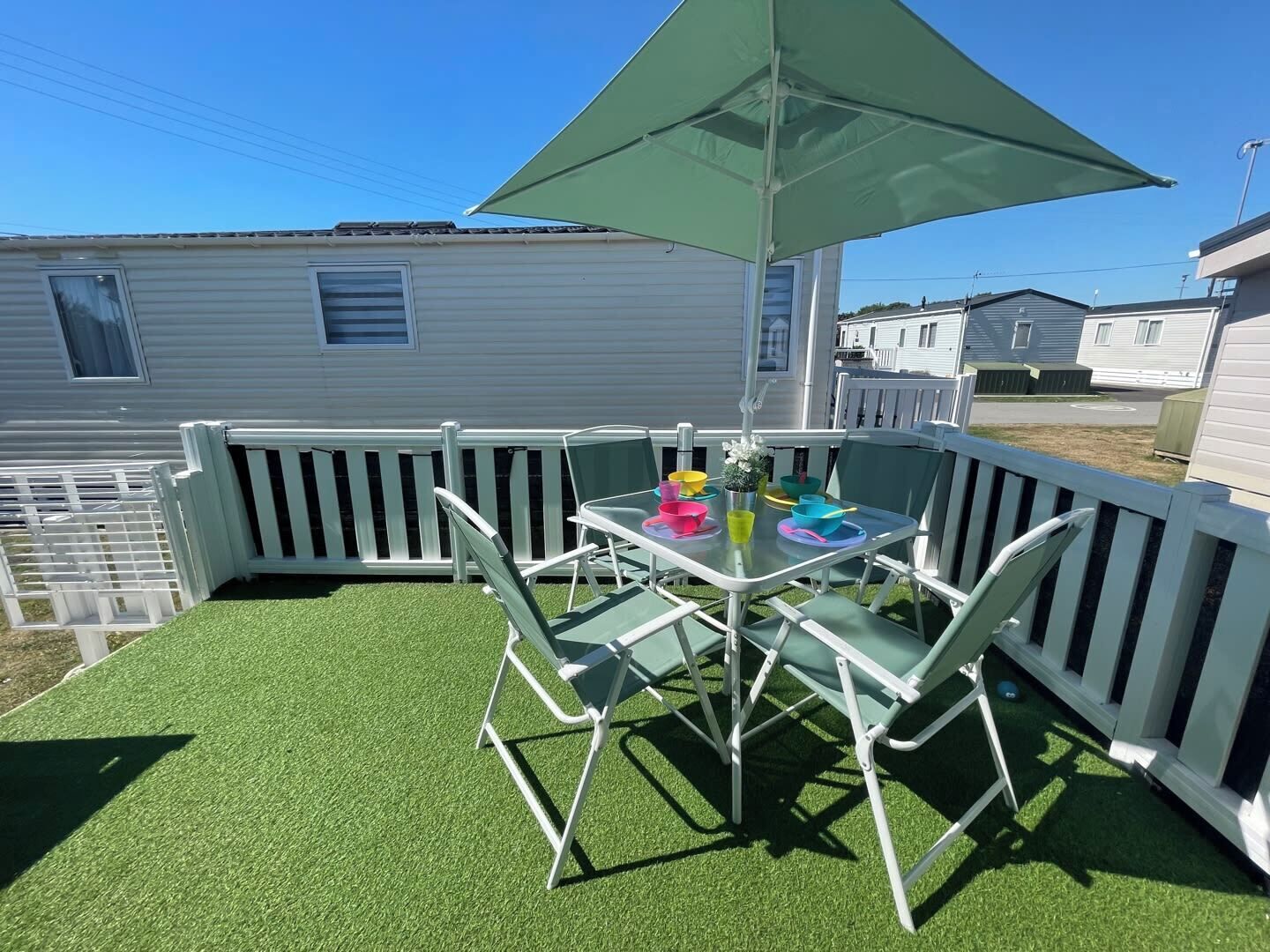 Un patio con mesa y sillas, rodeado por una cerca blanca, con casas móviles al fondo.