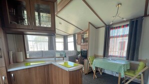 A compact RV kitchen with wooden cabinets, a small dining area with a checkered tablecloth, and a window with colorful curtains. - 27 Tower View Pevensey Bay Holiday Park Beach (Pevensey)