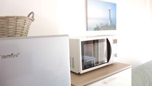 Un horno microondas sobre un gabinete blanco, una cesta de mimbre sobre un refrigerador blanco, y un cuadro en la pared.