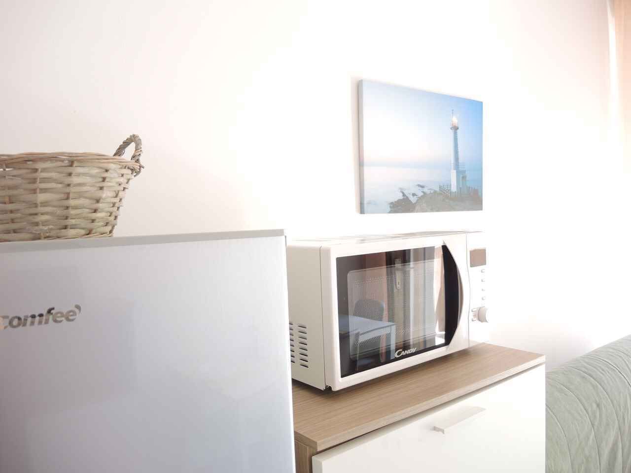 Un horno microondas sobre un gabinete blanco, una cesta de mimbre sobre un refrigerador blanco, y un cuadro en la pared.