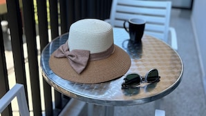 Una mesa redonda con un sombrero y gafas de sol encima, una silla blanca y una barandilla negra.