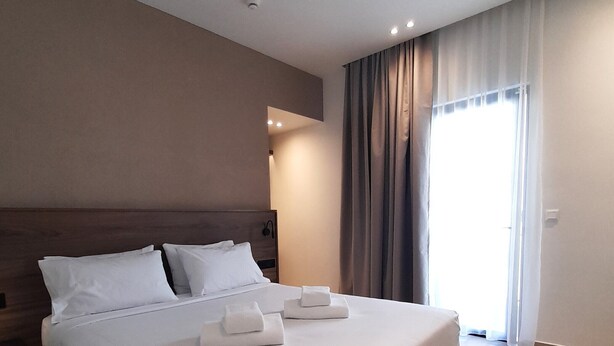 Una cama grande con ropa de cama blanca, cabecera de madera y dos almohadas. Hay dos toallas dobladas sobre la cama. La habitación tiene un ventanal con cortinas, una lámpara de techo y un despertador fijado a la pared.