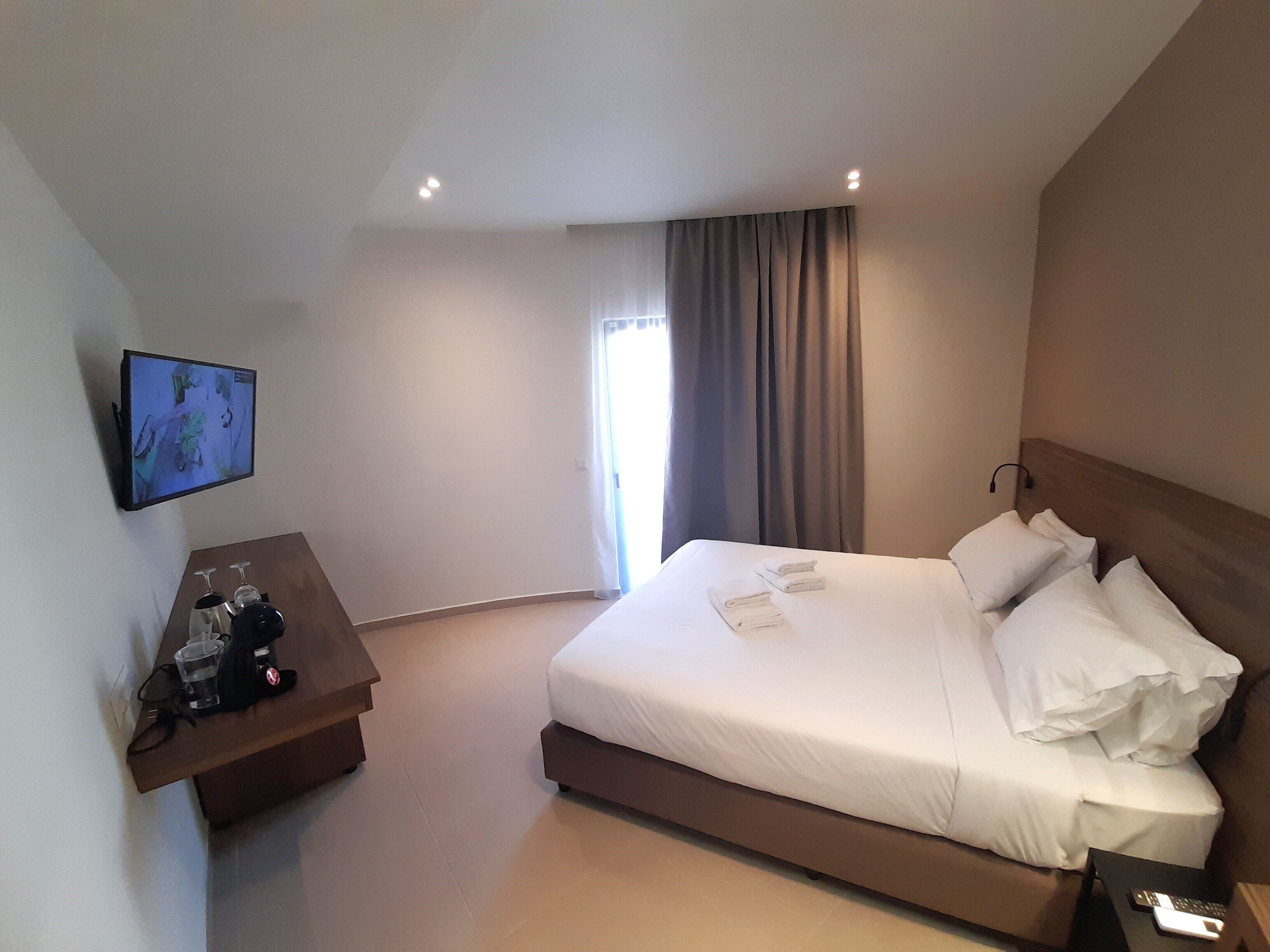 Habitación de hotel con una cama grande, un televisor montado en la pared, una mesita de noche con un teléfono y gafas, y una ventana con cortinas.