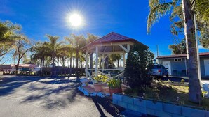 A covered outdoor area with a red roof, surrounded by palm trees and a paved road. - Seven Inn (Gumly Gumly)