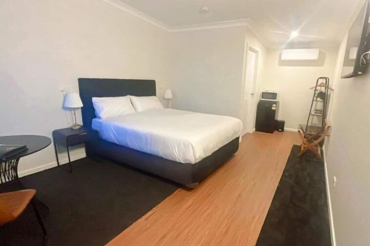 A hotel room with a bed, a desk, a chair, a small table, a TV, and a shoe rack.