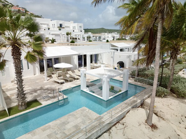 A pool area with lounge chairs and umbrellas, surrounded by palm trees and white buildings.