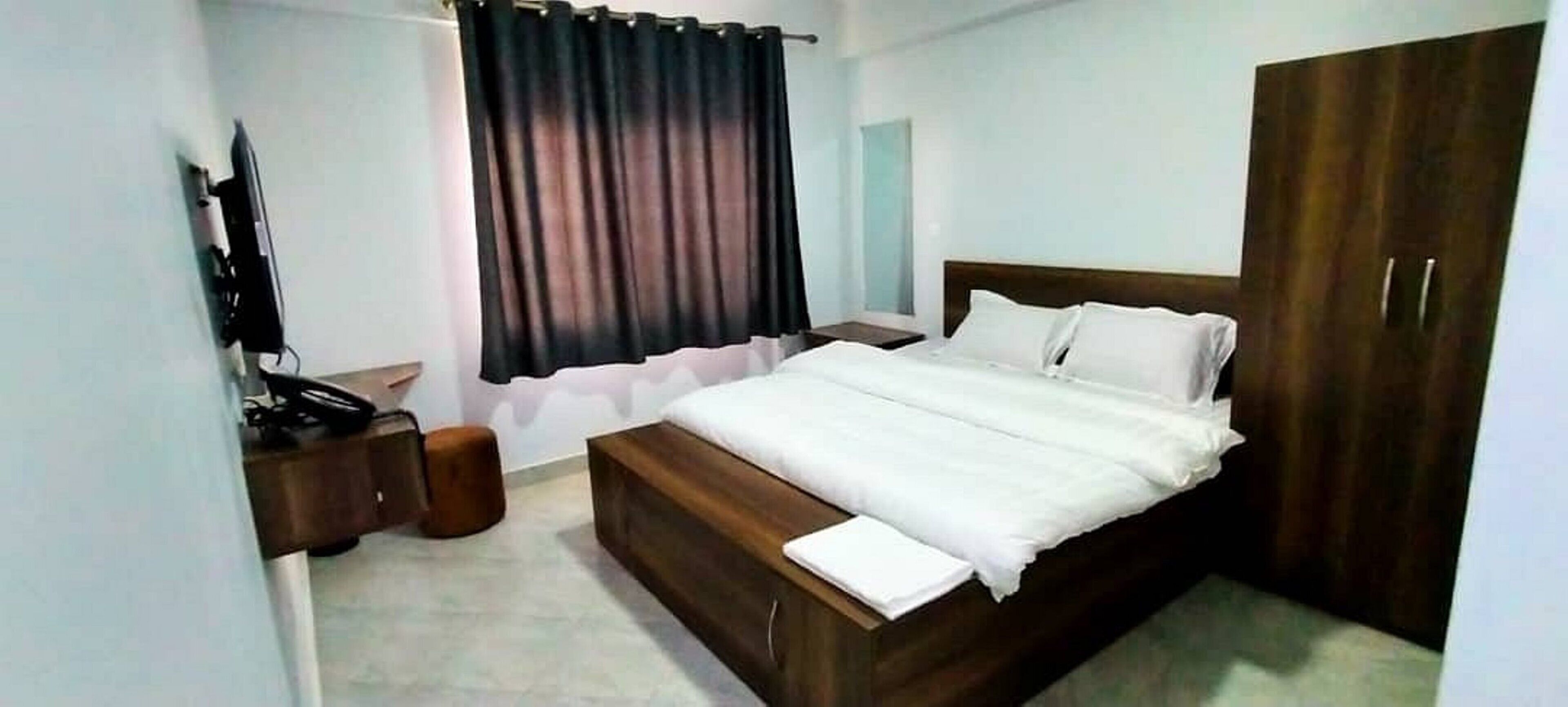 A bedroom with a wooden bed, white bedding, a nightstand, a TV, and a wardrobe.