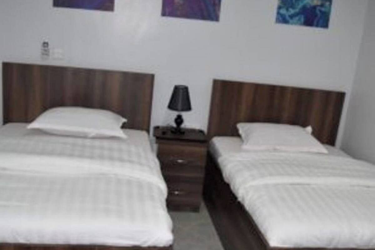 A hotel room with two beds, a nightstand with a lamp, and a small wooden stool.
