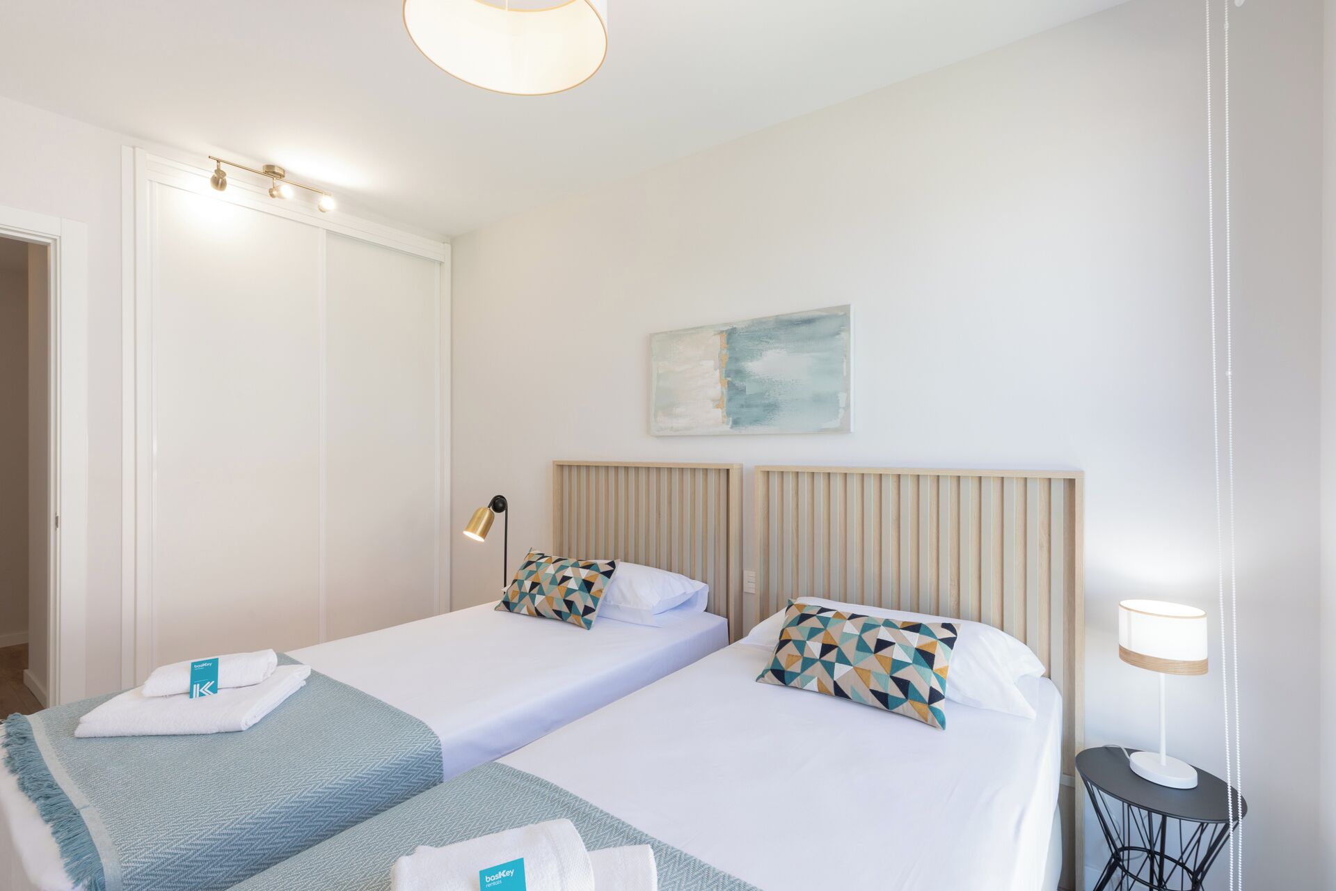A hotel room with two beds, a bedside table, a lamp, and a painting on the wall.