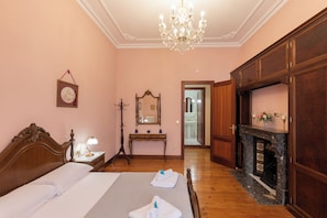Une chambre d’hôtel avec un grand lit, une armoire en bois, un miroir, un lustre et une cheminée.