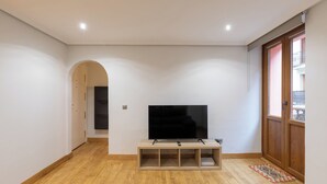 Een woonkamer met een flatscreen televisie op een houten standaard, een houten vloer en een groot raam met uitzicht op gebouwen.