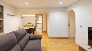 Een moderne woonkamer met een grijze bank, een eethoek met tafel en stoelen, en een keuken op de achtergrond.