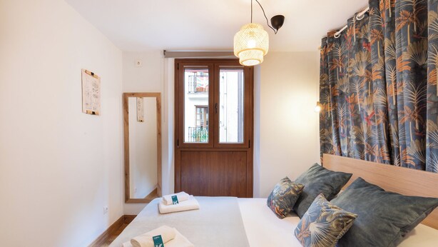 Een slaapkamer met een bed, kussens, een nachtkastje, een raam en een deur.