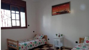 Una habitación con dos camas de madera, suelo a cuadros, una ventana con cortinas, una mesita de noche pequeña y blanca, y un cuadro en la pared.